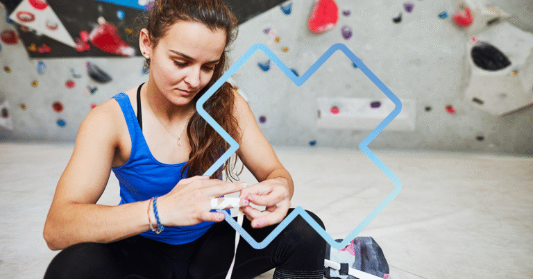 Femme assise dans un centre d’escalade, en train de mettre du ruban adhésif autour de son doigt blessé, avec un mur d’escalade en arrière-plan.